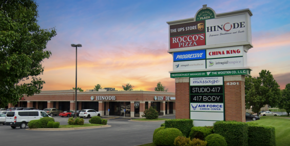 A detailed shot of the Aundria Plaza business directory sign in front of the shopping center at sunset. The sign lists businesses including The UPS Store, Rocco’s Pizza, Hinode, China King, Progressive, and more. Below the sign, manicured bushes and green grass surround the base. In the background, the shopping center features a brick exterior with arched walkways and parked cars. The sky displays a mix of warm sunset tones blending into cool blue.