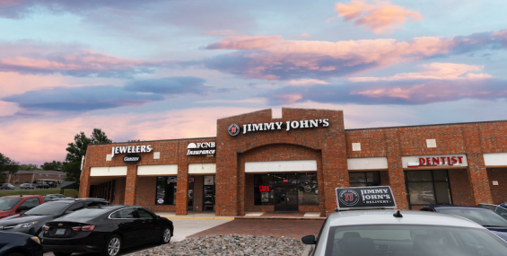 A brick strip mall features several businesses, including "Jimmy John’s," a jewelry store, an insurance office, and a dentist. A white Jimmy John’s delivery car is parked in front, along with several other vehicles. The parking lot has a decorative rock bed. The sky in the background is a blend of warm pink and blue colors at dusk.