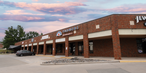 A brick shopping center with covered walkways and multiple storefronts, including "Farmers Insurance," is shown. A silver sedan is parked near the entrance, and additional businesses are visible in the distance. The sky is filled with pink and blue tones at sunset, casting a warm glow over the scene.