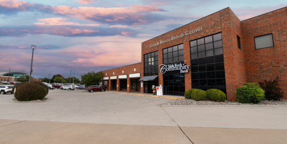 A brick commercial building with large glass windows houses "Table Rock Community Bank" and "Ozark Neuro Rehab Center." The parking lot is mostly empty, with a few cars visible in the distance. Landscaping includes trimmed bushes and rock beds. The sky is a mix of pink and blue hues, indicating sunset.