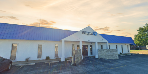 A single-story commercial building with a white exterior and a bright blue metal roof. The entrance features a triangular awning with the "Wilgus IQ" logo above the glass doors. The parking lot in front has a few spaces, with some construction materials leaning against the building. A large tree stands to the right, and the sky is a mix of golden hues, suggesting early morning or late afternoon.