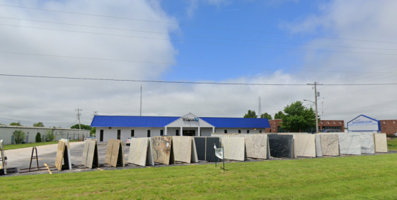 A white, single-story commercial building with a bright blue metal roof is set behind a display of large stone slabs arranged in rows on the asphalt. The sign above the main entrance reads “Wilgus IQ.” In the background to the right, another building with a sign reading “SHOWROOM” is visible. The foreground features a grassy area, and the sky above is partly cloudy with some patches of blue.