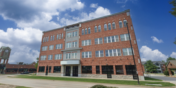 A four-story red brick building with arched windows and a symmetrical design, situated along a landscaped street. The ground floor features large glass storefronts, ideal for commercial leasing. The sky is bright blue with scattered clouds, and a nearby shopping center is visible in the background.