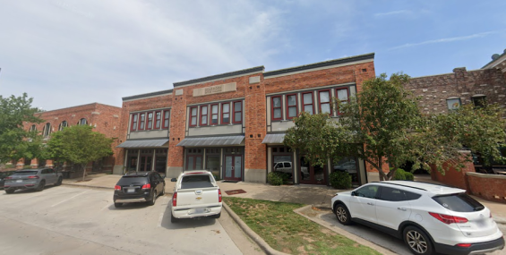 A two-story red brick office building with large windows and a classic architectural design. A sign above the entrance reads "Dearborn Office Building." The facade features metal awnings over the ground-floor entrances, and trees line the front. Several cars are parked along the curb.