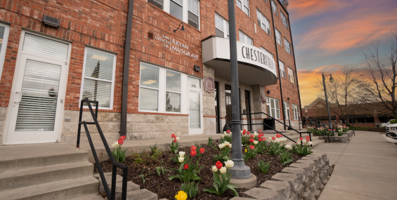 A close-up of the Chesterfield Lofts II entrance at sunset, framed by blooming red, white, and yellow tulips. The brick facade features a "Chesterfield" sign above double doors, with a law office sign for Bryan Musgrave to the left. The warm glow of the sunset enhances the inviting atmosphere.