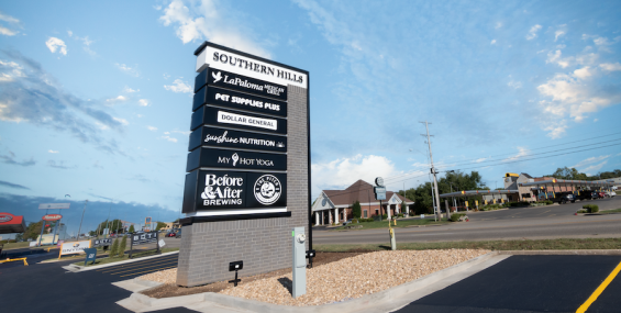 A tall, modern shopping plaza sign with a gray brick base and black panel sections, standing against a bright blue sky with scattered clouds. The sign displays various business names, including "La Paloma Mexican Grill," "Pet Supplies Plus," "Dollar General," "Sunshine Nutrition," "My Hot Yoga," "Before &amp; After Brewing," and "The Pitch Pizza &amp; Pub." The surrounding parking lot is freshly paved, and the background includes a street with commercial buildings and a gas station.