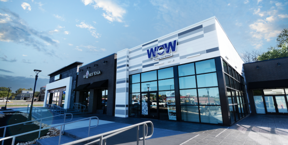 A close-up of two storefronts at Southern Hills Shopping Plaza, "My Hot Yoga" and "WOW - Watching Over Whiskers," a pet rescue organization. The buildings have a sleek design with black and white elements, large glass windows, and contrasting textures. A modern ramp with metal railings leads up to the entrances, ensuring accessibility. The sky is bright blue with a few wispy clouds, and reflections of nearby buildings and cars are visible in the glass windows.