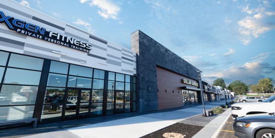 A row of contemporary retail stores at Southern Hills Shopping Plaza with large glass windows and modern exterior finishes. The visible businesses include "XGEN Fitness Private Personal Training" and "Dollar General." The building facade features a mix of white, gray, and dark stone materials, with a wooden accent section. The parking lot in front has several cars parked, and a landscaped sidewalk runs along the storefronts.