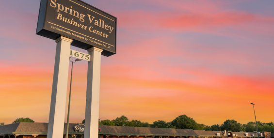 A tall, black and white sign reading "Spring Valley Business Center" stands against a colorful sunset sky. Below, the sign states that the center is professionally managed by The Wooten Co. LLC. The number "1675" is displayed on a separate panel beneath the main sign. In the background, a strip mall with brick storefronts and green trim lines an empty parking lot.