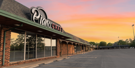 A close-up view of a store named "Piano Craft" in the Spring Valley Shopping Center. The store’s sign features a stylized script logo with "Piano" in white and "CRAFT" in bold red letters. The brick building has green trim and large reflective glass windows. The parking lot in front of the store is mostly empty, with a sunset sky reflecting in the windows.