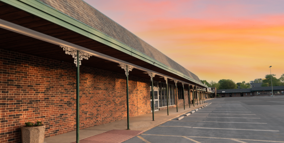 A row of covered walkways supported by green poles with decorative white brackets runs along the brick storefronts of the Spring Valley Shopping Center. The textured brick walls and green awning trim contrast with the soft hues of the sunset in the background. A red doormat sits in front of one of the entrances, and a large concrete planter with flowers adds a touch of greenery. The parking lot remains mostly empty.