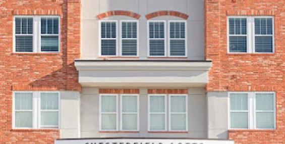 The front entrance of Chesterfield Lofts, a four-story brick building with large windows and a modern design. A sign above the entrance reads "Chesterfield Lofts," and the ground floor features commercial spaces with glass storefronts. The exterior combines red brick with stone accents, creating a contemporary yet classic aesthetic.