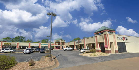 A slightly angled shot of Crossroads Shopping Center, showing various retail stores, including a restaurant with a visible "Hinode" sign. The architectural design features beige and red tones, decorative towers with green rooftops, and a parking lot with multiple vehicles. The sky is partly cloudy with blue patches visible.