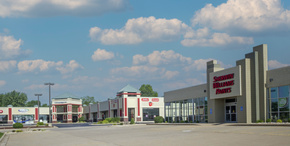 A wide-angle view of Crossroads Shopping Center in Nixa, Missouri, featuring a Sherwin-Williams Paints store in the foreground with large windows and a beige brick exterior. The shopping center extends into the background, showcasing multiple storefronts with red and beige accents, pyramid-shaped green rooftops, and a mostly empty parking lot under a bright blue sky with scattered clouds.