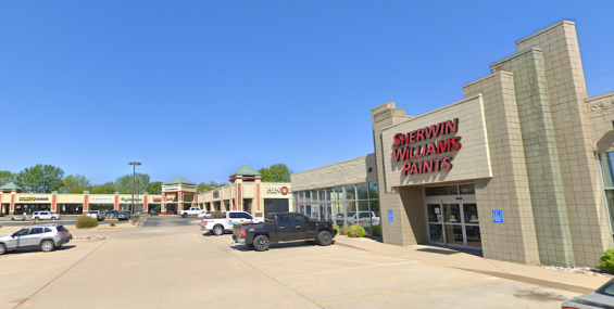 Another perspective of Crossroads Shopping Center, focusing on the Sherwin-Williams Paints store on the right side, with the rest of the shopping center extending into the distance. The parking lot has several vehicles, and the red and beige storefronts with green rooftops are clearly visible under a bright, cloudless blue sky.