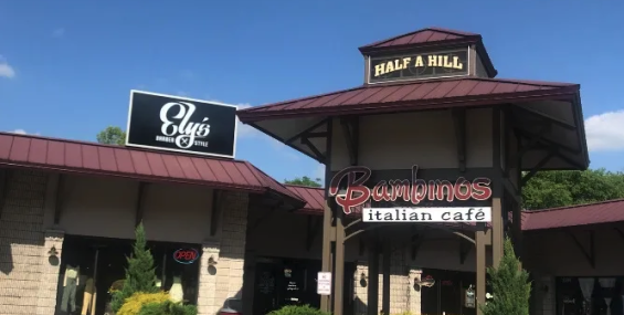 A section of Half A Hill Shopping Center featuring the entrance to Bambinos Italian Café, which has a distinctive black and red sign. Above the café, there is a wooden structure displaying the name "Half A Hill." To the left, Ely’s Barber &amp; Style has a black sign with white lettering. The buildings have a rustic design with stone walls and a red metal roof, and there is landscaping with small bushes and trees.