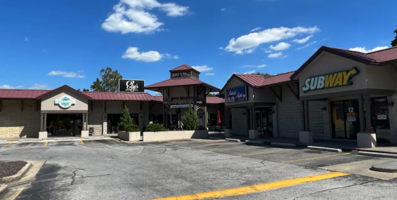 A wider view of Half A Hill Shopping Center, showing multiple storefronts, including Bambinos Italian Café, Ely’s Barber &amp; Style, a Subway restaurant, and another bakery. The buildings have a cohesive architectural style with stone exteriors, red metal roofs, and brown wooden accents. A parking lot with a yellow curb and a few empty spaces is in front of the shops, under a bright blue sky with scattered clouds.