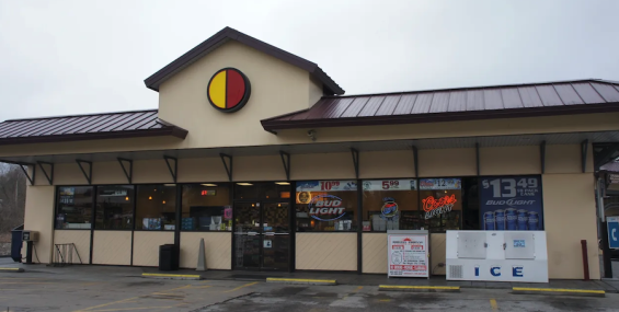 A convenience store with a beige exterior and a red and yellow circular logo above the entrance. The store has a brown metal roof and large front windows displaying beer advertisements. An ice machine sits outside near the entrance, and a parking lot with faded yellow lines is visible in the foreground.
