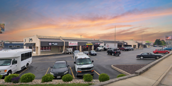 A shopping center with a row of beige, single-story retail buildings under a vibrant sunset sky. The storefronts include an esports gaming lounge, a donut shop, and other local businesses. The parking lot in front is active with various vehicles, including a shuttle bus, cars, and a delivery truck. Small green shrubs and a landscaped sidewalk frame the foreground.