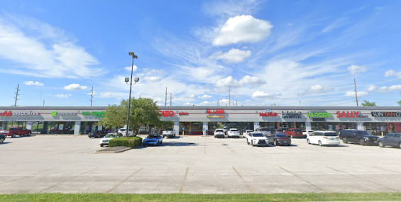 A wide-angle view of Nixa Center, a retail shopping plaza in Nixa, MO, featuring a single-story strip mall with multiple storefronts. Visible businesses include Murney Associates, H&amp;R Block, Pet Supplies Plus, Cato, El Lago Mexican Restaurant, Top Nails, Halo Hair Studio, Springfield Vapors, Sally Beauty, and Sun Tan City. The parking lot in front of the center has numerous cars parked, with a few trees and light poles scattered throughout. The sky is bright blue with scattered clouds, creating a clean and
