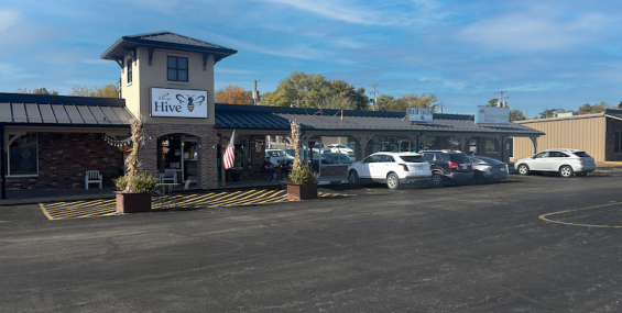 Exterior view of East Center in Willard, Missouri, showing a row of small businesses under a long covered walkway. The Hive storefront is centered beneath a small tower, featuring a white sign with a bee logo. Several planters and an American flag decorate the entrance. Multiple cars are parked in front of the shops, and the scene is set on a bright day with a blue sky.