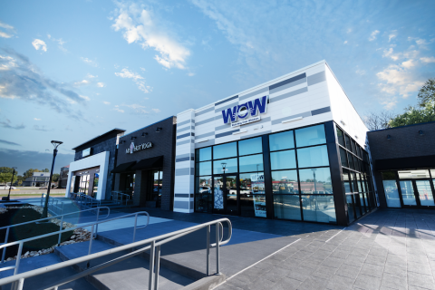 A close-up of two storefronts at Southern Hills Shopping Plaza, "My Hot Yoga" and "WOW - Watching Over Whiskers," a pet rescue organization. The buildings have a sleek design with black and white elements, large glass windows, and contrasting textures. A modern ramp with metal railings leads up to the entrances, ensuring accessibility. The sky is bright blue with a few wispy clouds, and reflections of nearby buildings and cars are visible in the glass windows.
