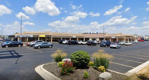 A shopping center with a row of tan-colored, single-story retail buildings under a bright blue sky with scattered white clouds. Visible businesses include a pharmacy, a chicken restaurant, and other retail stores. The large parking lot is partially filled with cars, and small, well-maintained shrubs and flowers line the edges of the lot. The shopping center appears well-maintained with clear signage.