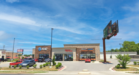 A modern shopping center with a stone and wood exterior, featuring AT&T, Missouri Title Loans, and Mercy GoHealth Urgent Care. The storefronts have large glass windows, with visible signage for each business. A parking lot with cars and neatly trimmed landscaping is in front of the building. A tall billboard stands to the right, and a Planet Fitness sign is visible in the background. The sky is bright blue with a few scattered clouds, and the overall setting appears well-maintained and inviting.