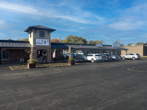 Exterior view of East Center in Willard, Missouri, showing a row of small businesses under a long covered walkway. The Hive storefront is centered beneath a small tower, featuring a white sign with a bee logo. Several planters and an American flag decorate the entrance. Multiple cars are parked in front of the shops, and the scene is set on a bright day with a blue sky.